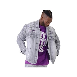 May include: A light gray denim jacket featuring distressed details and white and purple paint splatter effects. The jacket is worn over a purple t-shirt with white text that reads "FLY WITH US" in a bold, blocky, and slightly textured font.