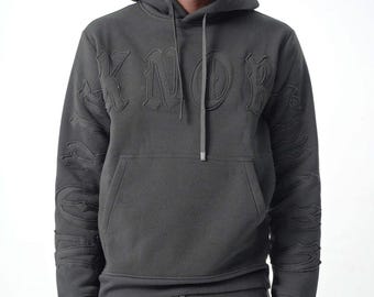May include: A black hooded sweatshirt featuring tonal, raised gothic-style lettering across the chest and down both sleeves. The garment includes a front kangaroo pocket, a drawstring hood with matching black cords, and ribbed cuffs.