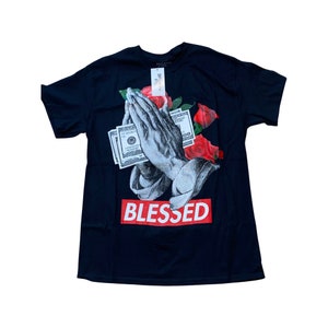 Könnte beinhalten: Schwarzes T-Shirt mit einem grafischen Druck von betenden Händen, die einen Geldschein mit roten Rosen halten. Der Text "BLESSED" ist in Rot unter dem Grafikdruck.