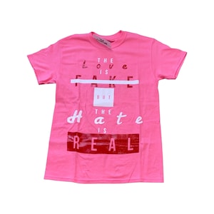 Könnte beinhalten: Pinkes T-Shirt mit dem Aufdruck "The love is fake but the hate is real" in Weiß und Rot.