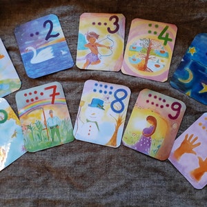 May include: Ten colorful cards with illustrations and braille dots representing the numbers 1 through 10. The cards are arranged in a semi-circle on a brown fabric surface.