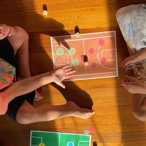 May include: Two children play a board game on a wooden floor. The game board is a basketball court with red, orange, and green circles. The children are using dice to move their pieces around the board.