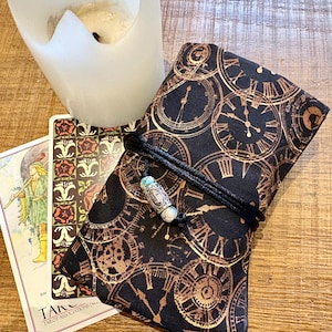 May include: A black tarot card bag with a gold clock face pattern, tied with a black cord and a decorative bead. Two tarot cards and a white candle are next to the bag. The cards have illustrations and text.