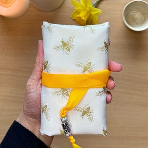 May include: A cream-coloured fabric pouch with a gold bee pattern, secured with a bright yellow ribbon. A decorative silver and bead charm hangs from the ribbon. The pouch is designed for storing small items.