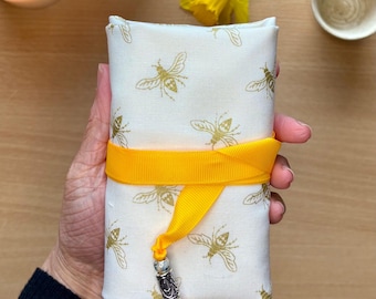 May include: A rectangular, cream-colored fabric pouch with a gold bee pattern, wrapped with a bright yellow ribbon. A decorative silver and bead charm hangs from the ribbon. The pouch is likely for storing small items.