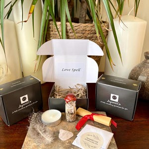 Peut inclure: Un kit 'Sortilège d'Amour' avec une petite bouteille en verre, une bougie, une plume, un cristal de quartz rose et un parchemin noué avec un ruban rouge. Le kit comprend deux boîtes noires avec le logo et le texte AyamiiCo.