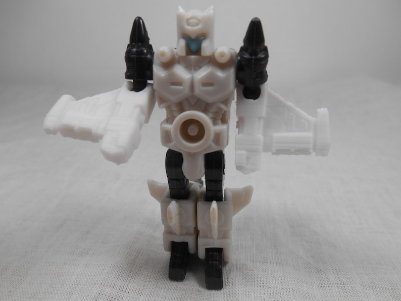 May include: A white and black action figure of a robot with wings. The robot has a white body with black accents on the shoulders and legs. The robot's head is white with a blue face. The figure is standing with its arms outstretched.