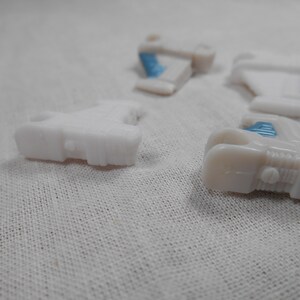 May include: Several small, white plastic toy car parts are scattered on a white fabric surface. Some parts have small blue accents. The pieces appear to be from a larger toy vehicle, possibly a car or truck.