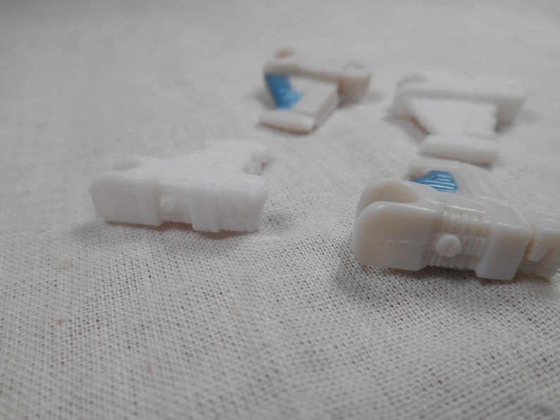 May include: Several small, white plastic toy car parts are scattered on a white fabric surface. Some parts have small blue accents. The pieces appear to be from a larger toy vehicle, possibly a car or truck.