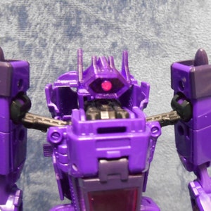 May include: A close-up of a purple robot toy with a red eye and black details. The toy has a boxy design with articulated arms and legs. The toy's head has a distinctive shape with a central eye and two protrusions.
