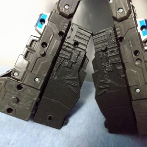 May include: Close-up of two black plastic robot legs with intricate panel detailing. The legs are connected by a hinge and feature silver screws and small blue and white accents. The overall design suggests a mechanical or robotic theme.