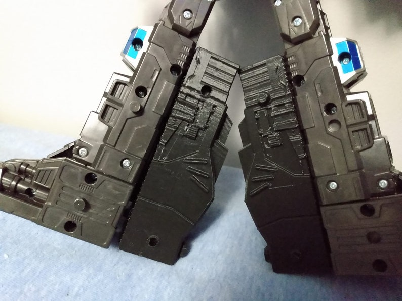 May include: Close-up of two black plastic robot legs with intricate panel detailing. The legs are connected by a hinge and feature silver screws and small blue and white accents. The overall design suggests a mechanical or robotic theme.