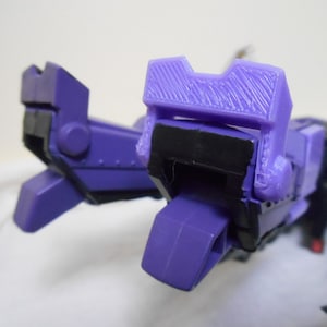 May include: A close-up of a purple and black toy robot. The robot has a boxy design with articulated joints and a textured surface on the head. The toy is made of plastic and has a glossy finish.
