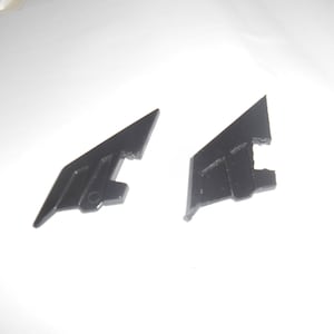May include: Two black plastic components, each with a triangular form and a rectangular base. They feature parallel lines and a small hole. The items are set against a white backdrop.