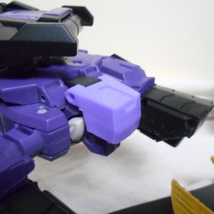Peut inclure: Gros plan d'un robot jouet violet et noir. Le robot présente un design détaillé avec des formes angulaires et une finition mate. L'image met en évidence les détails mécaniques du jouet.