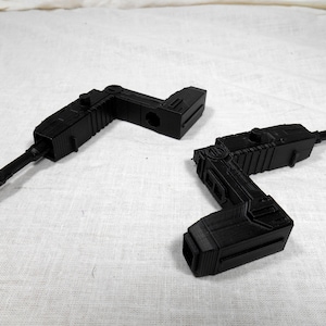 May include: Two black, angular, 3D-printed objects, possibly components of a larger device, are displayed against a white background. Each object has a distinct L-shape with a cylindrical extension. The objects appear to be made of plastic.