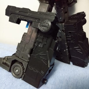 May include: A black, articulated robot toy, featuring intricate paneling and a wheel detail on the lower leg. The toy's design includes angular shapes and mechanical elements, suggesting a transforming robot.