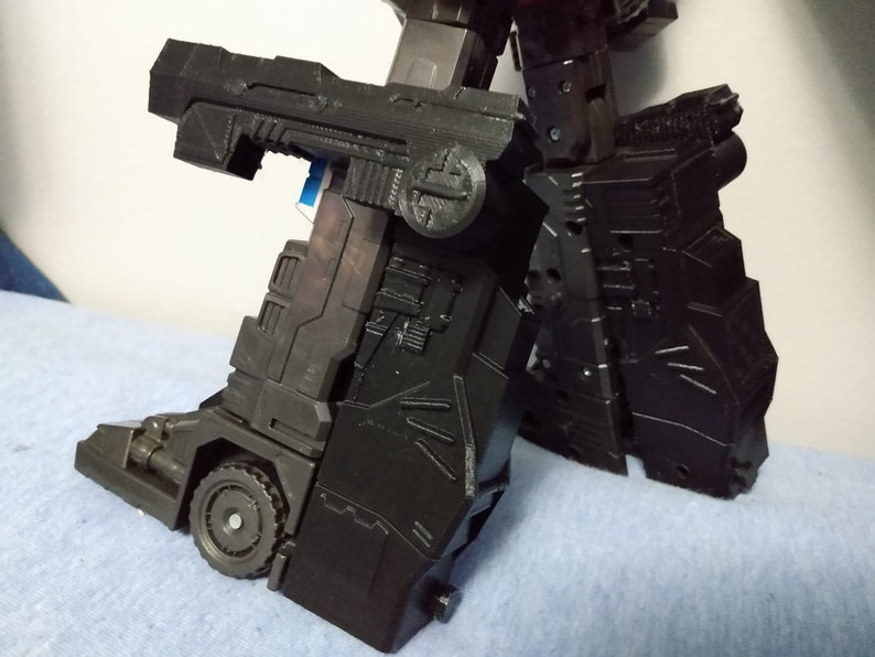 May include: A black, articulated robot toy, featuring intricate paneling and a wheel detail on the lower leg. The toy's design includes angular shapes and mechanical elements, suggesting a transforming robot.