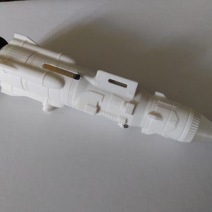 Può includere: Modello bianco stampato in 3D di un'astronave o di un razzo. Il modello ha una base circolare nera e un'estremità anteriore affusolata. Presenta elementi di superficie dettagliati, tra cui componenti rettangolari e cilindrici.