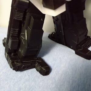 May include: Close-up of the lower portion of a black robot figure. The image focuses on the feet and lower legs, showcasing intricate geometric designs and a textured surface. The figure is set against a light blue background.