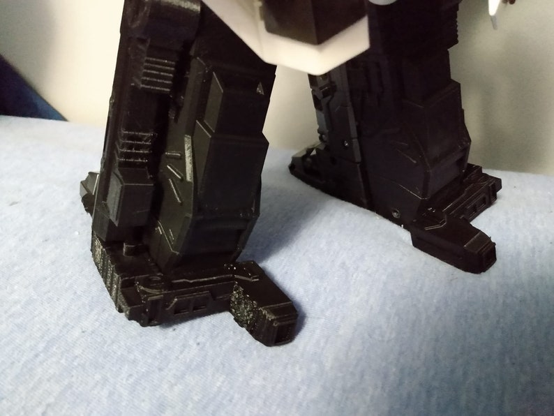 May include: Close-up of the lower portion of a black robot figure. The image focuses on the feet and lower legs, showcasing intricate geometric designs and a textured surface. The figure is set against a light blue background.