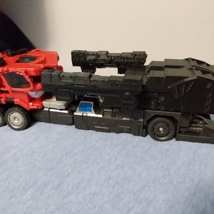 May include: A red and black toy truck with multiple wheels. The front cab is red with silver and white accents. The back section is black with a textured design. The toy truck is on a blue surface.