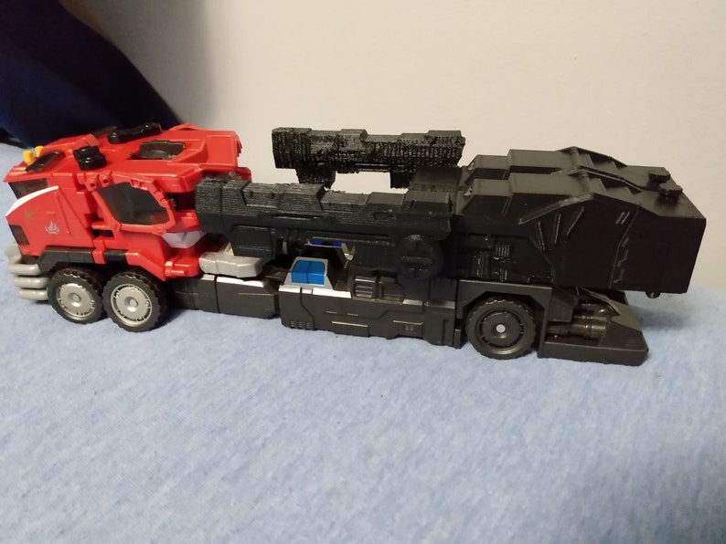 May include: A red and black toy truck with multiple wheels. The front cab is red with silver and white accents. The back section is black with a textured design. The toy truck is on a blue surface.