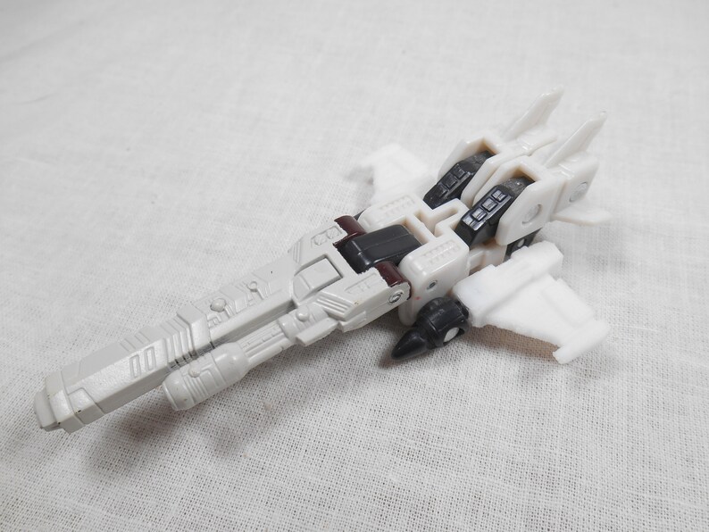 May include: A white and gray toy airplane with black and maroon accents. The toy has a long, cylindrical body with wings and a detailed design. The toy is made of plastic and appears to be a model or collectible item.