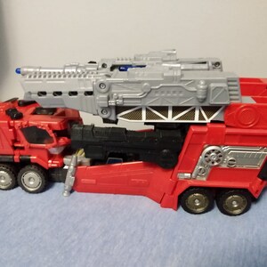 May include: A red and gray toy truck with multiple wheels and a large gray structure on top. The truck has a detailed design with silver accents and a ladder-like component. The toy is likely a fire truck or a similar emergency vehicle.