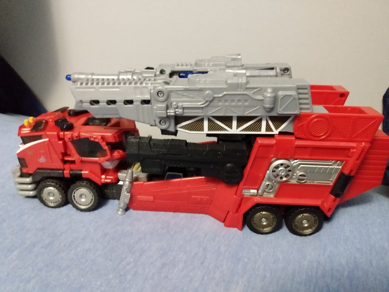 May include: A red and gray toy truck with multiple wheels and a large gray structure on top. The truck has a detailed design with silver accents and a ladder-like component. The toy is likely a fire truck or a similar emergency vehicle.