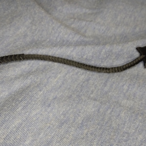 Può includere: Un oggetto a forma di freccia grigio scuro con un fusto strutturato e segmentato e una punta di freccia appuntita. La freccia è su una superficie di tessuto blu chiaro. Il design suggerisce un oggetto decorativo o simbolico.