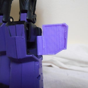 May include: A close-up of a purple and black toy robot's leg. The leg is mainly purple with black details. A purple panel is attached to the leg, featuring a textured design.