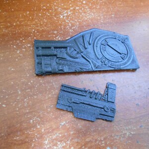 May include: Two black, intricately designed, 3D-printed components. The larger piece features a circular design element and detailed mechanical features. The smaller piece has a similar aesthetic, with a focus on mechanical details. Both are set against a wooden surface.