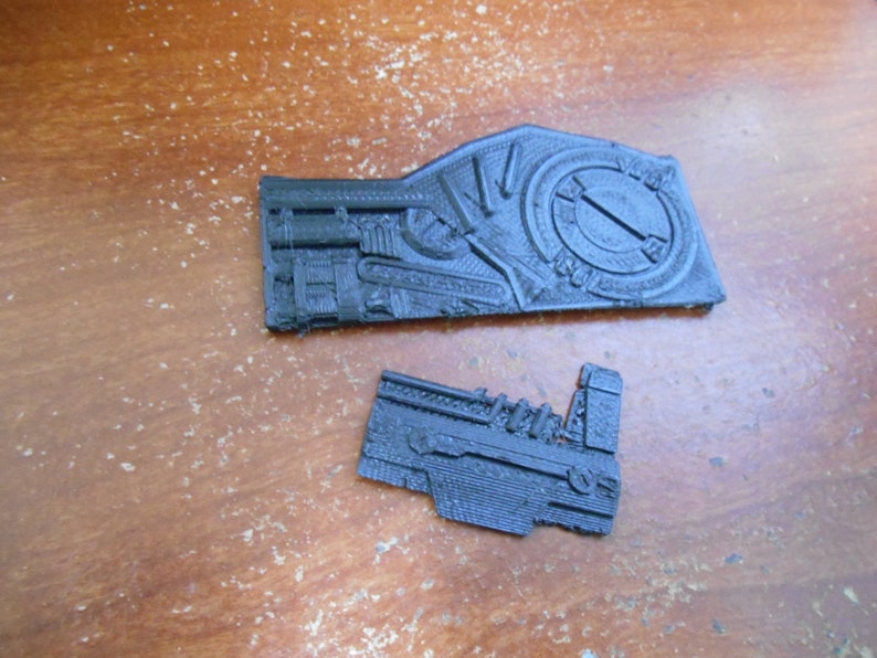 May include: Two black, intricately designed, 3D-printed components. The larger piece features a circular design element and detailed mechanical features. The smaller piece has a similar aesthetic, with a focus on mechanical details. Both are set against a wooden surface.