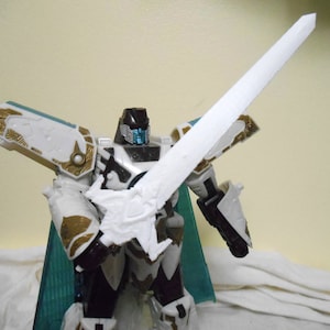 Puede incluir: Figura de acción de robot blanco y dorado sosteniendo una gran espada blanca. El robot tiene una cabeza negra con detalles azules y una armadura blanca con detalles dorados. Se ve una capa turquesa.