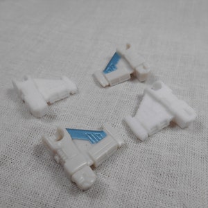 May include: Four small, white plastic toy airplane parts with light blue accents. The parts are scattered on a white fabric surface. The toy parts have a wing-like shape and are likely components of a larger model.