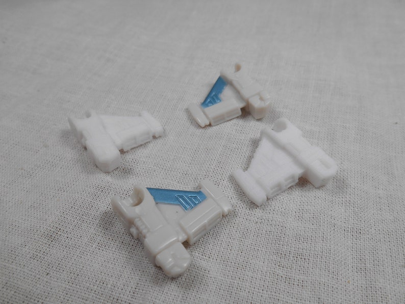 May include: Four small, white plastic toy airplane parts with light blue accents. The parts are scattered on a white fabric surface. The toy parts have a wing-like shape and are likely components of a larger model.