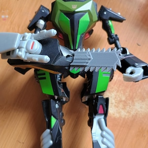 May include: A black and green robot action figure featuring a metallic green chest plate with a small gold insignia, articulated limbs, and a large gray chainsaw attachment held in one hand. The figure has detailed mechanical joints and molded parts.
