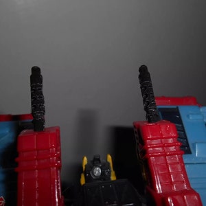 Puede incluir: Primer plano de un robot de juguete azul, rojo, amarillo y negro. El juguete tiene dos estructuras verticales negras y texturizadas. El dise&ntilde;o del robot presenta una combinaci&oacute;n de formas geom&eacute;tricas y detalles mec&aacute;nicos, lo que sugiere un juguete transformable.