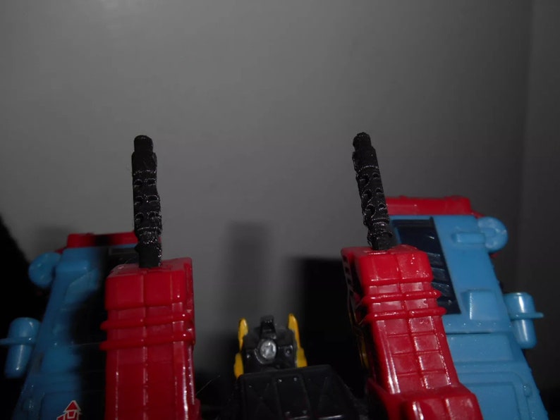 Puede incluir: Primer plano de un robot de juguete azul, rojo, amarillo y negro. El juguete tiene dos estructuras verticales negras y texturizadas. El dise&ntilde;o del robot presenta una combinaci&oacute;n de formas geom&eacute;tricas y detalles mec&aacute;nicos, lo que sugiere un juguete transformable.