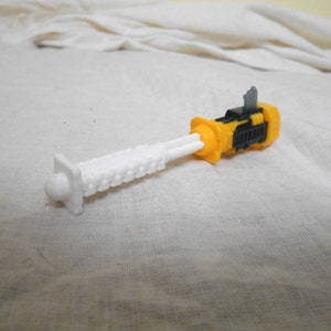 Może przedstawiać: Zabawka transformator w kolorze ż&oacute;łtym, czarnym i białym. Zabawka ma biały, teksturowany element przypominający ostrze oraz ż&oacute;łto-czarny korpus. Zabawka leży na białej powierzchni.