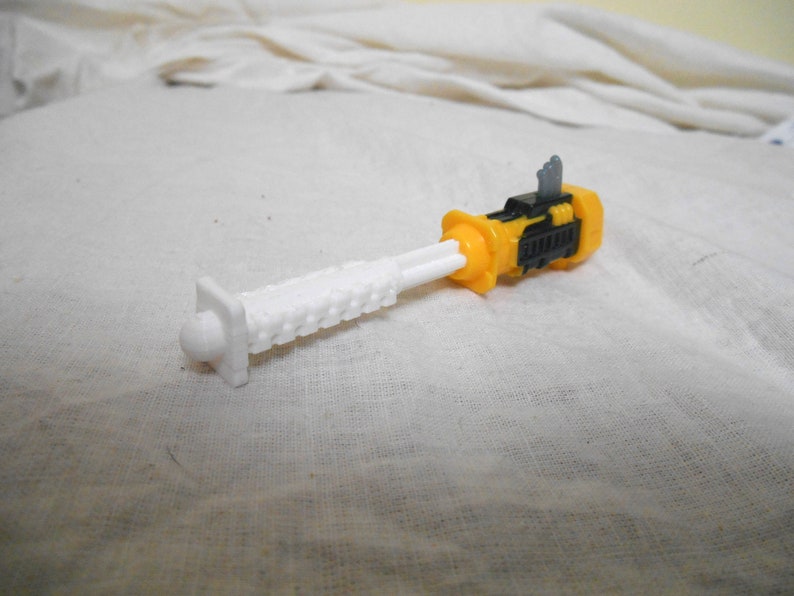 Może przedstawiać: Zabawka transformator w kolorze ż&oacute;łtym, czarnym i białym. Zabawka ma biały, teksturowany element przypominający ostrze oraz ż&oacute;łto-czarny korpus. Zabawka leży na białej powierzchni.