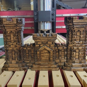 May include: A detailed wooden castle model with a dark brown finish. The model features two towers and a central structure, showcasing intricate carved details. It is positioned on a wooden base, with a CNC machine in the background.