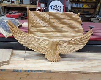 Può includere: Una scultura in legno di un'aquila calva americana con le ali spiegate, che tiene una bandiera americana negli artigli. L'aquila e la bandiera sono scolpite in uno stile realistico, con dettagli intricati. La scultura è realizzata in legno chiaro e ha una finitura naturale.