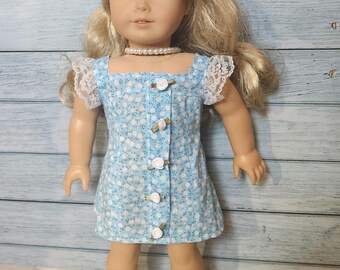May include: A doll wearing a light blue floral print dress with white lace sleeves and white rosebud buttons. The doll has blonde hair and is wearing a pearl necklace and white shoes. The dress is a square neck style.