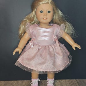 May include: A pink dress with a lace overlay and a satin bodice with a criss-cross ribbon detail. The dress is worn by a doll with blonde hair and green eyes.