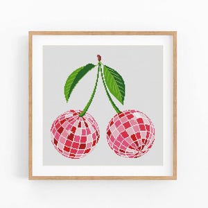 May include: A framed cross-stitch artwork featuring two cherries. The cherries are rendered in shades of pink and red, with a geometric, disco ball-like pattern. Green leaves and stems connect the cherries. The artwork is set against a light gray background.