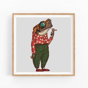 May include: A cross-stitch pattern featuring a cartoon frog wearing a red and white polka dot shirt, green pants, and holding a cigarette.