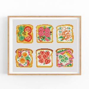 May include: A framed cross-stitch art piece featuring six different types of toast. Each toast slice is adorned with various toppings, including vegetables, meats, and fruits. The artwork is set against a white background and framed in a light brown wooden frame.