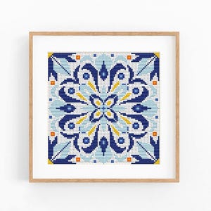 May include: A square cross-stitch embroidery featuring a symmetrical geometric pattern in shades of dark blue, light blue, white, and yellow, enclosed within a light-coloured wooden frame. The design displays a central floral-like motif.